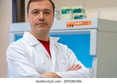 Portrait Of Scientist Mask In Room With Laboratory Box In UV Light. Happy Successful Male Worker Of Scientific Laboratory Standing By Laboratory Shelf Inside Office In Front Of Camera