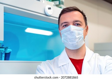Portrait Of Scientist In Medical Mask In Room With Laboratory Box In UV Light. Tired Successful Male Worker Of Scientific Laboratory Sitting By Laboratory Shelf Inside Office In Front Of Camera