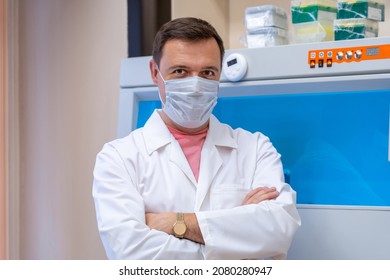Portrait Of Scientist In Medical Mask In Room With Laboratory Box In UV Light. Tired Successful Male Doctor Standing By Laboratory Shelf Inside Office In Front Of Camera