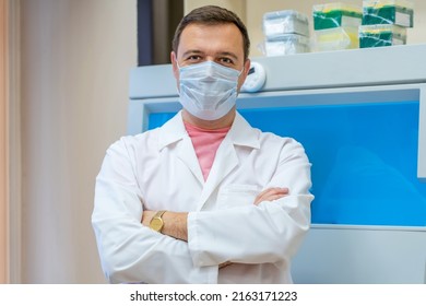 Portrait Of Scientist In Medical Mask In Room With Laboratory Box In UV Light. Happy Successful Male Doctor Standing By Laboratory Shelf Inside Office In Front Of Camera