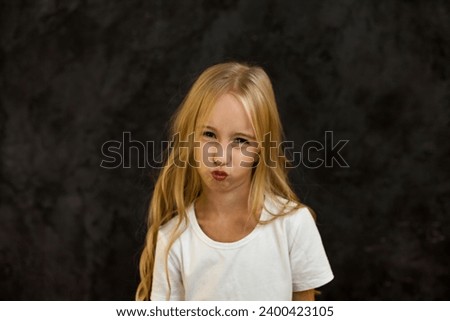 Portrait of scowl cover girl kid model in white t-shirt expression emotion, frowning looking at camera. Angry child 6 year old posing at black, studio shot. Kids emotional concept. Copy ad text space
