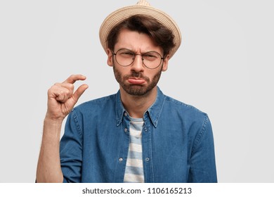 Portrait Of Scrupulous Bearded Young Male Shows Something Very Tiny With Hand, Has Upset Expression, Purses Lower Lip, Dressed In Denim Shirt With Straw Hat, Isolated On White Background. Too Small!