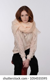 Portrait of a seated red-haired girl on a light gray background. She is dressed in an elegant lace blouse and a light scarf around her neck. Warm colors, gold ornaments. Fashion.