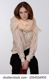Portrait of a seated red-haired girl on a light gray background. She is dressed in an elegant lace blouse and a light scarf around her neck. Warm colors, gold ornaments. Fashion.