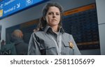 Portrait of Security Officer in Airport Terminal: Female TSA Worker in Uniform Looking at Camera. Diverse Crowd of Passengers Looking at Arrival and Departure Information Display in the Background.