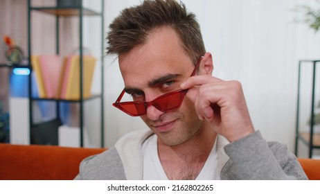 Portrait Of Seductive Cheerful Stylish Successful Man In T-shirt Wearing Red Sunglasses, Charming Smile, Blinking, Winking Eye, Flirting. Young Lovely Adult Guy Sitting At Home In Living Room On Couch