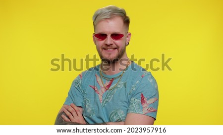Portrait of seductive cheerful tourist stylish man in blue t-shirt wearing sunglasses, charming smile. People emotions concept. Young lovely adult guy posing isolated on yellow studio wall background