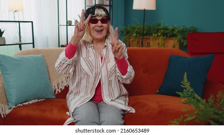 Portrait Of Seductive Stylish Caucasian Senior Old Grandmother In Black Cap Hat With Marijuana Wearing Red Sunglasses, Looking At Camera With Charming Smile. Elderly Modern Youngster Woman At Home