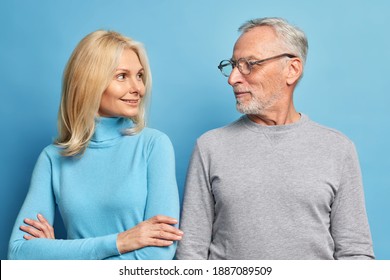 Portrait Of Self Confident Blonde Woman Stands Crossed Arms And Looks Happily At Her Mature Husband Have Pleasant Talk Together Look At Each Other Isolated Over Blue Background. Elderly Couple