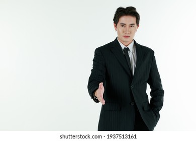 Portrait Of Self-confidence Young And Handsome Asian Businessman In Black Suit Standing In Elegant Pose And Give Forward Hand For Shake, Copy Space Studio Shot Isolated On White Background.