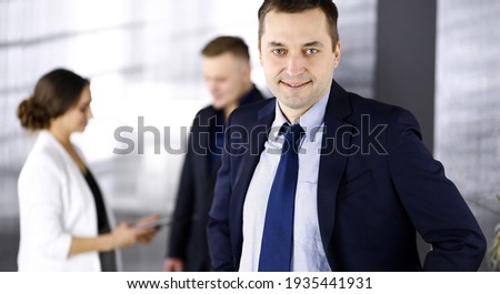 Portrait of a self-confident middle aged businessman in a blue suit, standing in a modern office with his colleagues at the background. Concept of business success