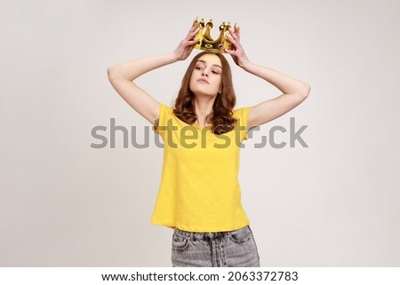 Portrait of selfish brown haired teenage girl in yellow T-shirt holding gold crown over head, having arrogance expression, privileged status. Indoor studio shot isolated on gray background.