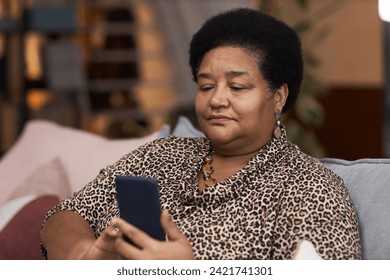 Portrait of senior African American woman holding smartphone and tapping screen using internet while relaxing at home - Powered by Shutterstock