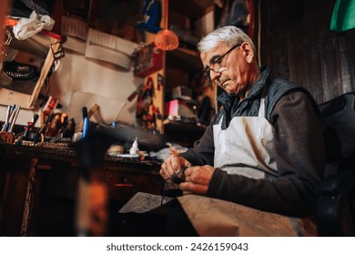 Portrait of a senior artisan sitting at cobbler's workshop and cutting leather parts for shoe making. An old shoemaker in apron is sitting at his workshop and preparing leather parts for shoe making. - Powered by Shutterstock