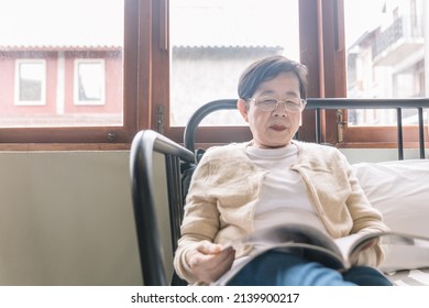 Portrait of senior Asian woman wearing sweater sitting on the sofa and enjoying reading a magazine at home.