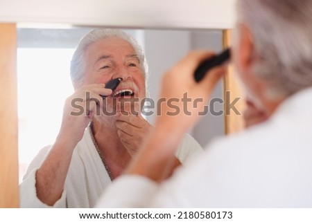 Portrait of senior bearded man in bathrobe while remove nose ear hair with help of little comfy trimmer looking at himself in the mirror