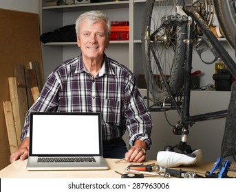 Portrait Of Senior Bike Shop Owner Sitting At Desk Behind The Laptop With White Screen. Active Old Man Looking At Camera And Smiling. Small Business. 