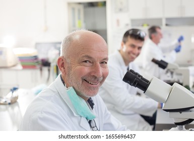 Portrait Of Senior Biologist Working On Microscope With Colleagues In Laboratory