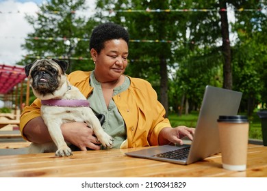 Portrait Of Senior Black Woman Using Laptop In Outdoor Cafe With Cute Pug Dog