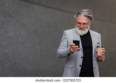 Portrait of a senior business executive man smiling and using mobile phone and drinking coffee. A elderly businessman standing on the street surrounded by buildings and using smartphone. Copy space. - Shutterstock ID 2227301723