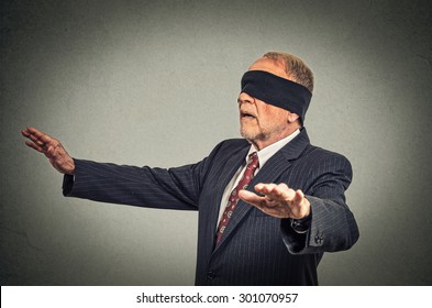 Portrait senior business man in suit blindfolded stretching his arms out isolated on gray wall background