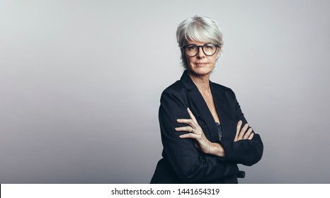 Portrait Of A Senior Businesswoman Looking At Camera. Senior Woman Entrepreneur Standing Against Grey Background With Her Arms Crossed.