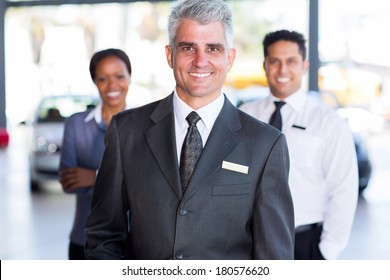 Portrait Of Senior Car Dealership Principal With Staff On Background