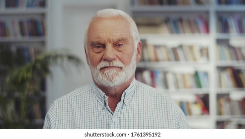 Portrait Of The Senior Caucasian Good Looking Grey-haired Man Lokking At The Side And Then Straight To The Camera With Serious Or Angry Face In The Library. Close Up.