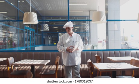 Portrait Of Senior Chef In White Uniform Walking In Empty Cafe Waiting For Customers. Restaurant Chef Checking Cleanliness Of Furniture Before Opening. Small Business Concept