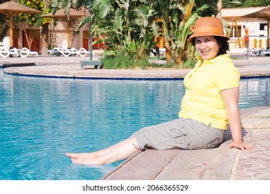 Portrait Of Senior Citizen Brunette Woman Sitting In Wicker Hat Sits On The Edge Of The Pool. Woman Put Her Feet In The Pool And Checks The Water Temperature. Recreation At The Resort