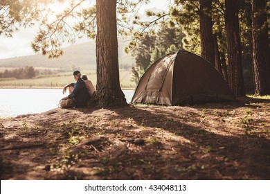 Portrait Of Senior Couple Camping By A Lake. Man And Woman Sitting Under A Tree Looking Away A The Lake On Summer Day.