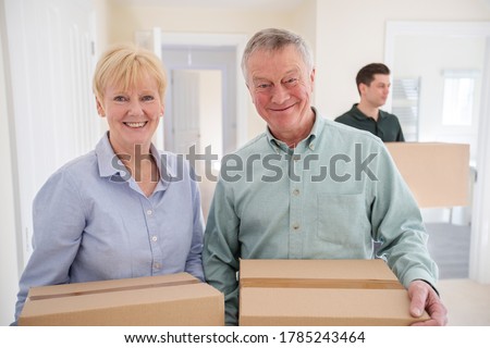 Portrait Of Senior Couple Downsizing In Retirement Carrying Boxes Into New Home On Moving Day With Removal Man Helping