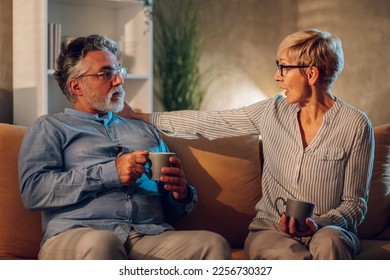 Portrait of a senior couple drinking coffee while sitting on a sofa at home and looking at each other while having a serious conversation. Modern interior. Copy space. - Shutterstock ID 2256730327