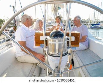 Portrait Of A Senior Couple With Friends Seated In A Sailing Boat Clinking Wine Glasses As Seen Through The Steering Wheel.