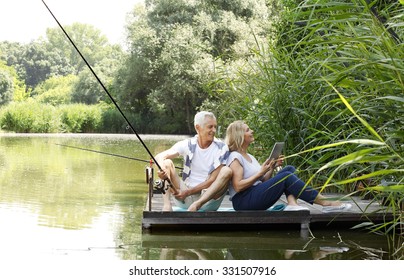 Portrait Of Senior Couple Relaxing At Lakeside. Old Man Fishing And Elderly Woman Using Digital Tablet While Sitting At Pier. 