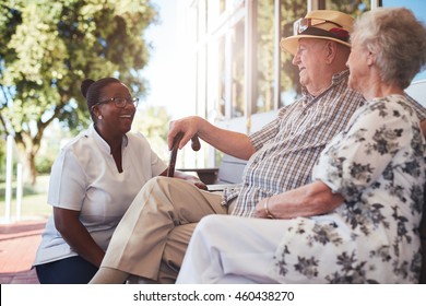 Portrait Of Senior Couple Relaxing On A Bench Outside Their Retirement Home. Caucasian Elderly Man And Woman Sitting With Female Nurse At Old Age Home.