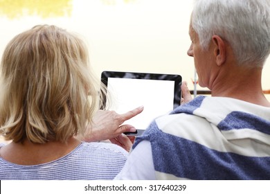 Portrait Of Senior Couple Sitting At Lakeside. Elderly Woman Holding Hand White Blank Screen Digital Tablet While Old Man Sitting Next To Her And Looking The Display.