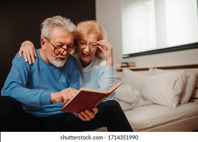 Portrait of senior couple sitting on bed in bedroom and reading from book with red covers. - Powered by Shutterstock