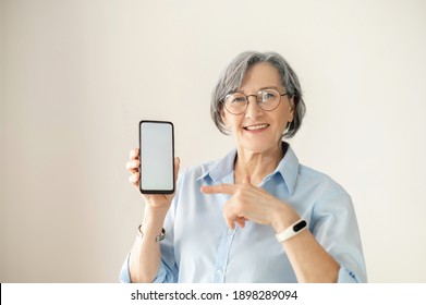 Portrait Of A Senior Elder Smiling Woman Showing Or Presenting Mobile Phone Application And Pointing Finger At The Smartphone Screen On Hand, Mature Businesswoman Isolated Over Gray Background