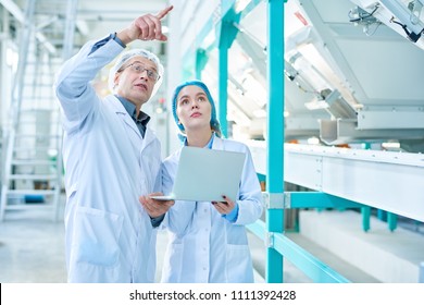 Portrait Of Senior Factory Worker Wearing Lab Coat Explaining Rules To Female Trainee And Pointing Up While Standing In Clean Production Workshop Of Modern Plant, Copy Space