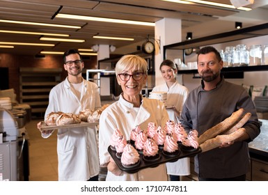 Portrait Of Senior Female Baker Holding Cupcakes In Her Hands At Bakery With Her Multi Generation Colleagues In Backgrounds.