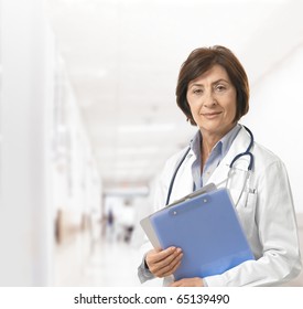 Portrait Of Senior Female Doctor On Hospital Corridor Holding Clipboard Looking At Camera Smiling.?