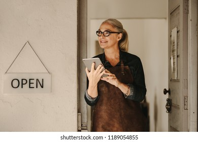 Portrait of senior female goldsmith standing at workshop entrance with digital tablet. Female jeweler wearing apron and holding a tablet pc at workshop door. - Powered by Shutterstock