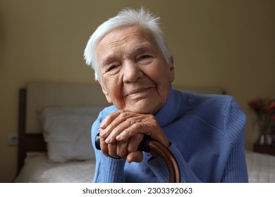 Portrait of a senior female sitting on the bed and leaning on a cane. Elderly woman resting her head on a handle of a walking cane. Close up, copy space for text, background. - Powered by Shutterstock