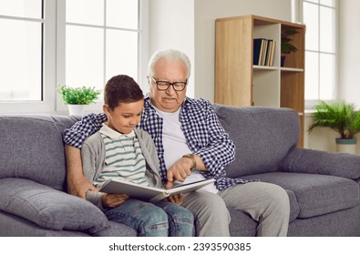 Portrait of a senior grandfather reading a book to a little smiling child boy in the living room or looking through a photo album sitting on sofa at home. Family leisure and generations concept. - Powered by Shutterstock