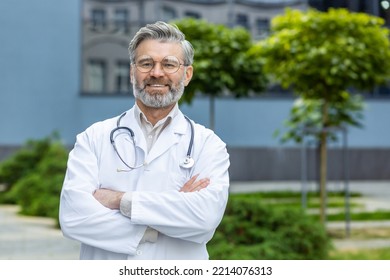 Portrait Of Senior Gray-haired Doctor, Mature Man In White Medical Coat With Crossed Arms And Stethoscope Smiling And Looking At Camera, Head Doctor Outside Modern Hospital