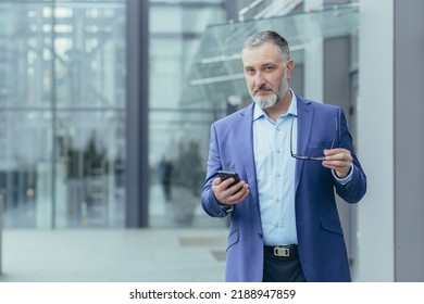 Portrait Of Senior Gray-haired Financier, Man Outside Office Building Holding Phone And Looking At Camera, Businessman Walking