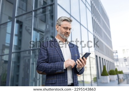 Portrait of a senior gray-haired male doctor standing in a suit outside the office and using a mobile phone, reading the news, typing a message, waiting for an appointment.