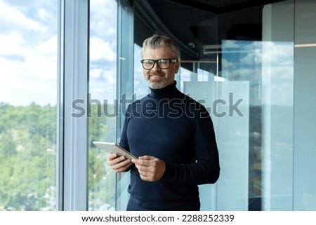 Portrait of a senior handsome gray-haired businessman in glasses standing in the office of a skyscraper by the window, holding a tablet in his hands and looking at the camera with a smile.
