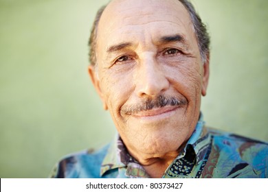 Portrait Of Senior Hispanic Man With Mustache Looking At Camera Against Green Wall And Smiling. Horizontal Shape, Copy Space